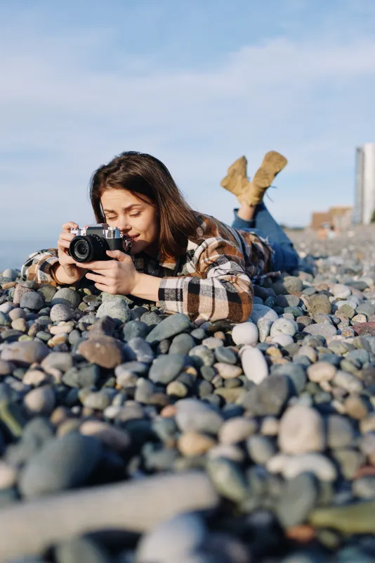 photographer woman beach rocks outdoor casual camera lens focus lying on pebbles capturing a sunlit seascape with concentration and artistic framing