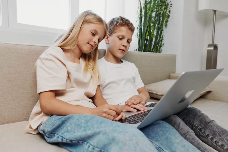 Two children sitting on a couch using a laptop together in a bright living room. Focused girl and boy collaborate on a project, concept for learning, technology, and teamwork at home.