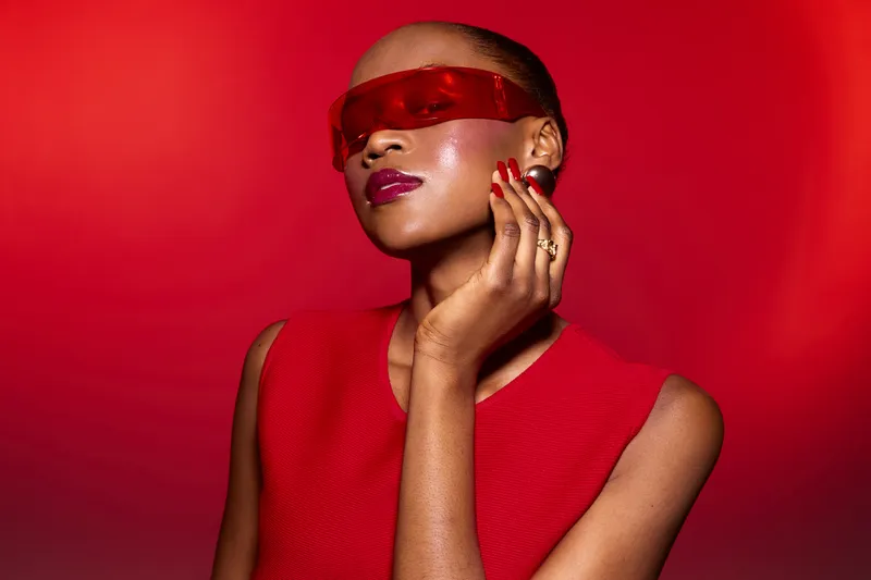 Stylish African American woman with dark skin wearing red futuristic sunglasses and sleeveless red dress poses confidently against red background. Concept of fashion, beauty, and modern style.