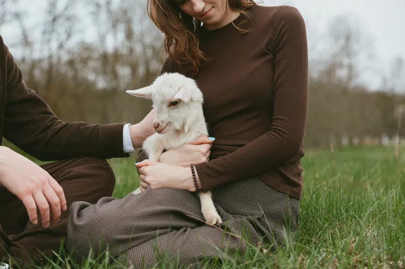 Lifestyle scene outdoors with a couple and a goat kid, calm moment on grass, gentle interaction, warm tones, candid expression, relaxed posture, natural light, rustic countryside mood