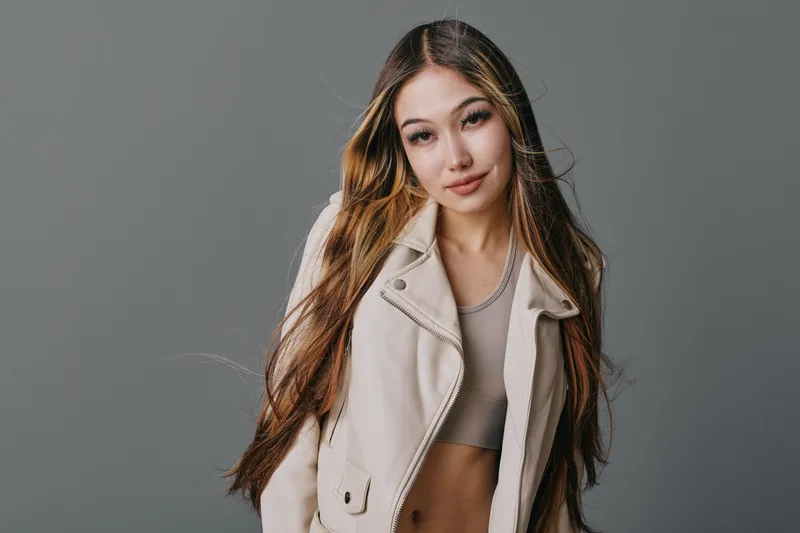 Young woman with long hair wearing cropped top and light jacket posing elegantly against gray background. Casual fashion style with natural makeup and confident expression.