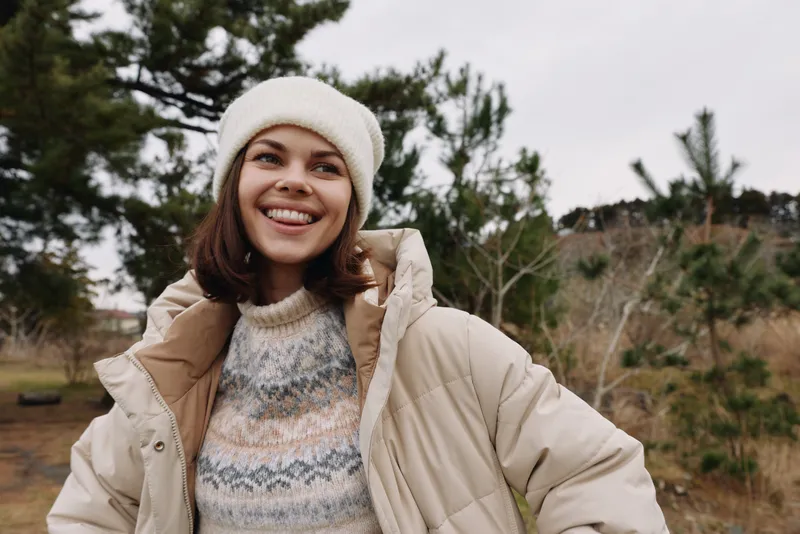 Smiling woman in a cozy winter outfit outdoors, wearing a puffy jacket, knit sweater and beanie, enjoying crisp weather among trees and natural scenery today.
