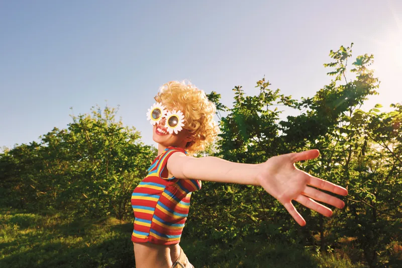 Woman in a striped swimsuit with flowers for eyes extends her arm in a sunny outdoor scene, retro film color, playful and carefree mood, vibrant summer vibe