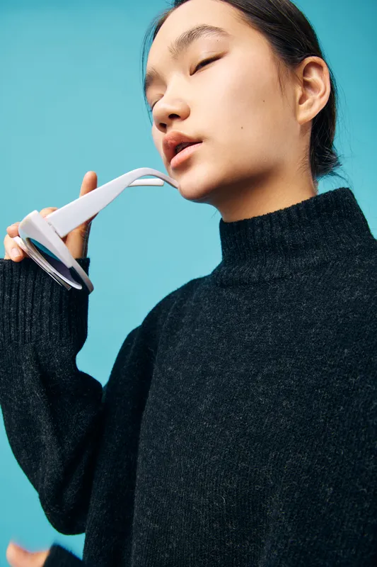 Young woman in black sweater holds white sunglasses near face with closed eyes and calm expression against blue background for fashion portrait or style concept.