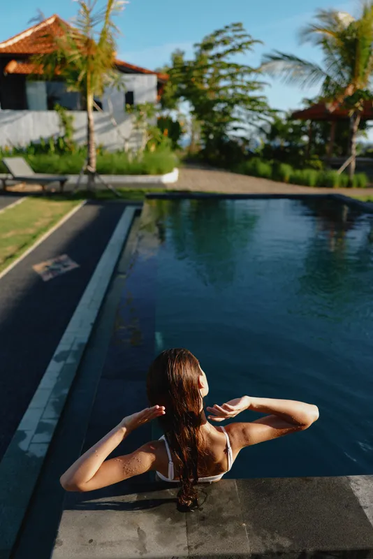 A woman relaxes at a sunny poolside edge with her back to the camera, hair braided. A tranquil lifestyle moment by water, capturing leisure, vacation and wellness vibes.