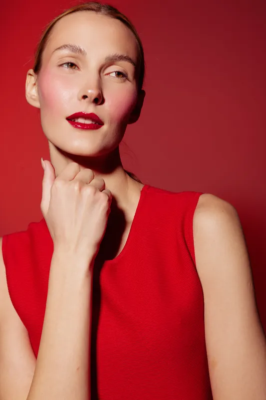 Young woman with red lipstick and red sleeveless top poses against red background. Fashion portrait with soft lighting highlights natural skin texture and confident expression.