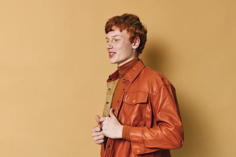 Young redhead man wearing orange leather jacket and turtleneck standing against beige background looking sideways with confident expression and relaxed posture.