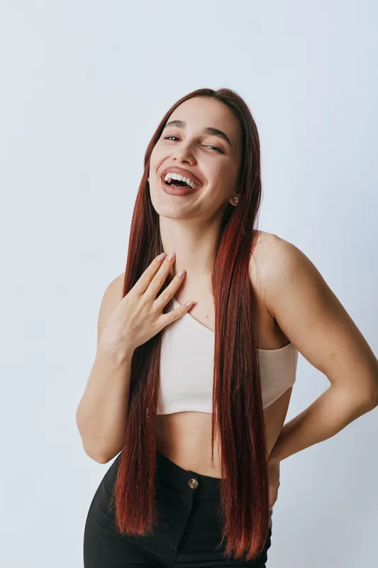Young woman with long straight hair laughing and posing in casual outfit against white background. Female model smiling with hand on chest and relaxed posture in studio.