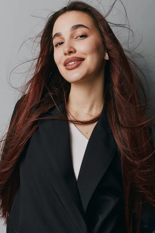 Young woman with long hair and confident smile wearing black jacket and white shirt standing against gray background with natural makeup and casual hairstyle for portrait.