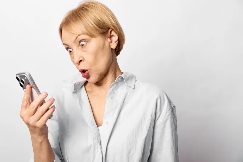 Surprised older woman holding a smartphone in a studio with white background, shocked eyes and open mouth, sudden communication reaction to a message