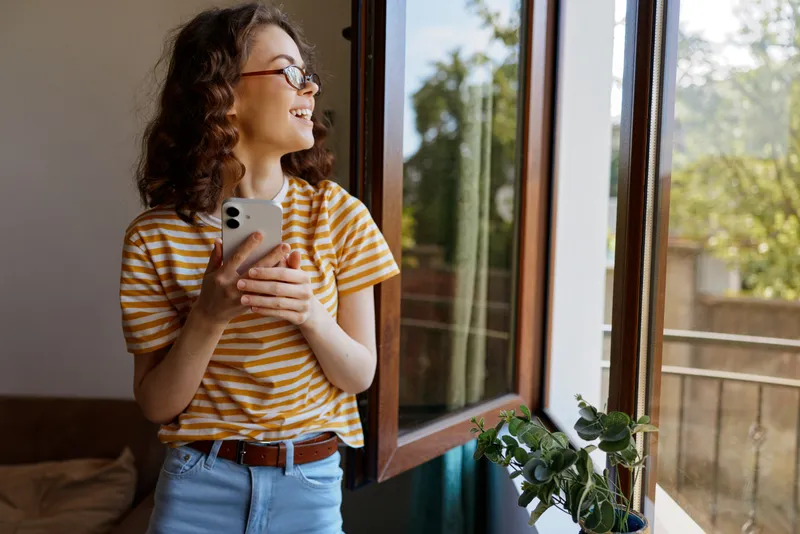 Cheerful woman indoors by a sunlit window, holding a smartphone, casual striped top, relaxed posture, warm natural light, home ambiance, greenery visible outside, lifestyle moment