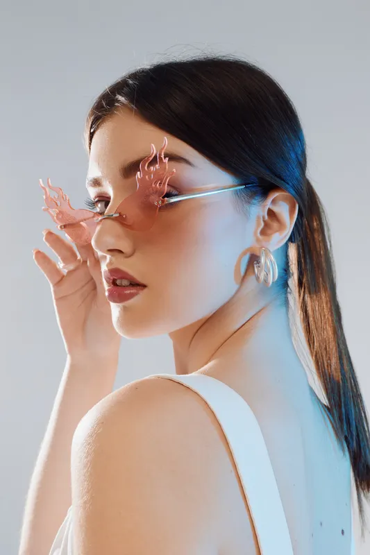 Young woman with long dark hair wearing unique pink sunglasses and gold earrings poses in a white top. Studio portrait with soft lighting. Concept of woman beauty fashion.