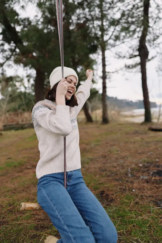 Girl on a swing outdoors in a knit sweater and beanie, smiling as she sways in a park setting with trees and wide open space, casual recreation and carefree moment captured in natural light.