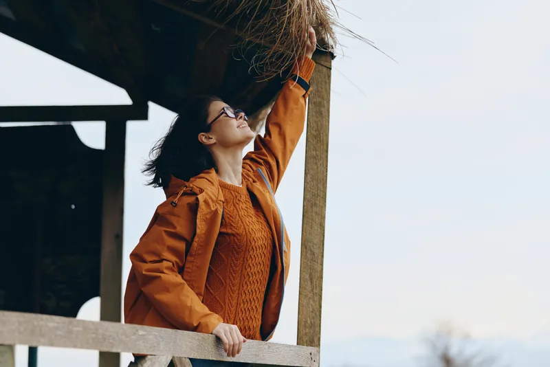 Woman lifestyle outdoors, smiling softly, wearing brown coat and sunglasses, leaning on wooden railing, sunlit windy air, relaxed posture, blurred nature background, casual travel mood