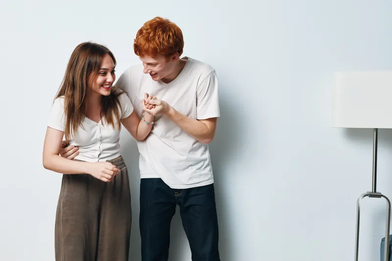Young couple smiling and laughing together standing near white wall with lamp, casual clothing and friendly interaction indoor for lifestyle and relationship.