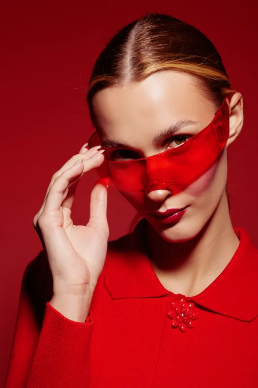 Young woman in red outfit and red translucent sunglasses poses against red background. Fashion portrait with sleek hairstyle and confident expression. Concept of style and modern accessories.