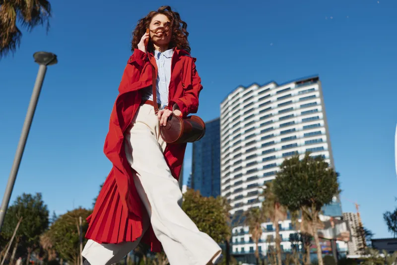 Confident woman in a red coat walks briskly through a sunny city street, white pants fluttering, modern buildings rise behind, wind tousling her curly hair.
