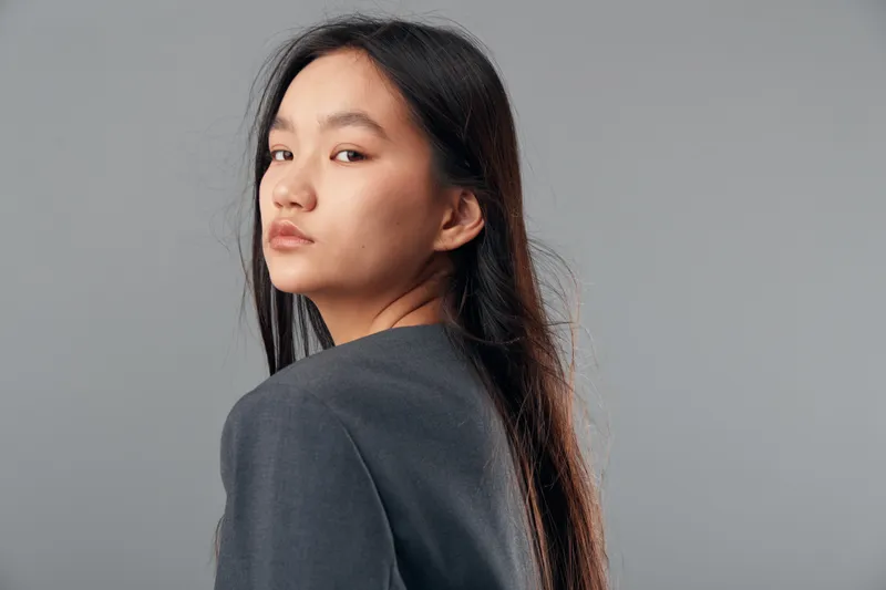 Portrait of young woman with long black hair and gray top looking sideways against plain gray background. Asian female model with serious expression and natural makeup in studio lighting.