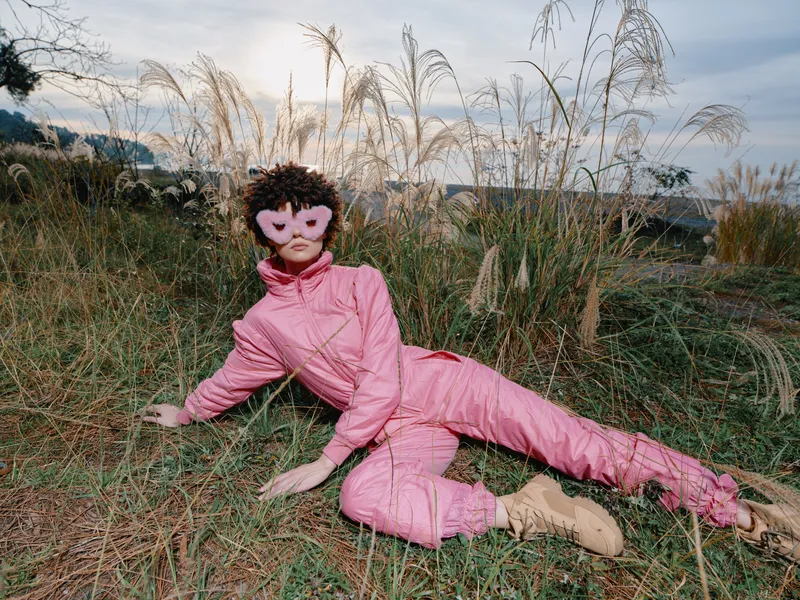 Fashion model in pink jumpsuit reclining in tall grass wearing a pink heart mask, outdoor landscape, editorial style with calm natural light and relaxed pose.
