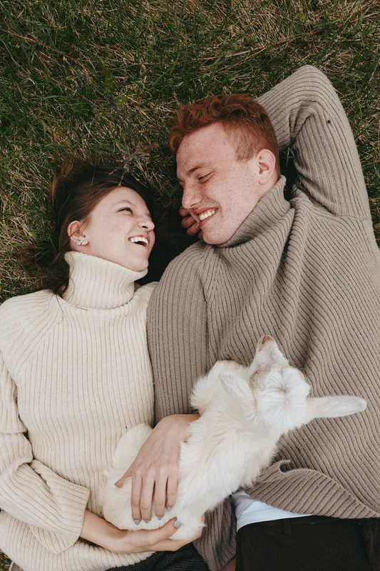 Joyful couple lying on grass in knitwear, sharing warm smiles and cuddling a fluffy white dog, creating a bright lifestyle moment of affection, relaxation, and outdoor happiness in a natural setting