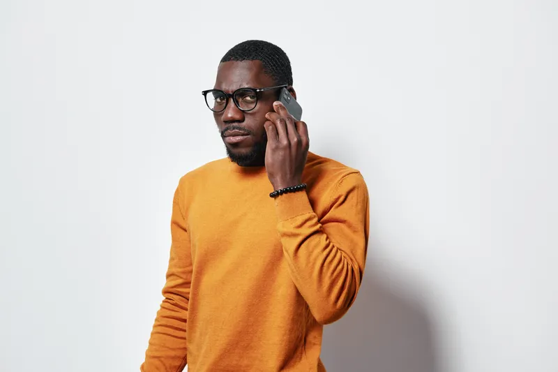 African American man with dark skin wearing glasses and orange sweater talking on smartphone against white background. Serious expression, communication and technology concept.