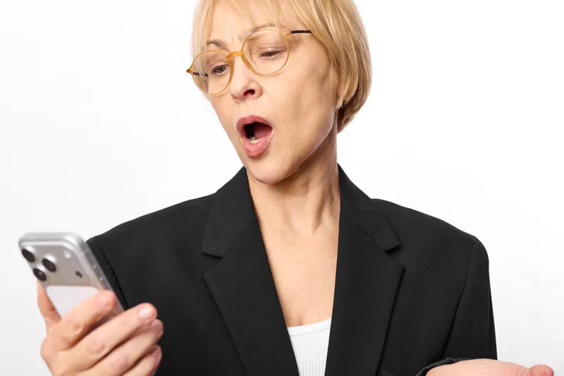 Senior woman in business suit with glasses, surprised expression while holding a smartphone, looking at screen, studio white background, communication moment, focused posture