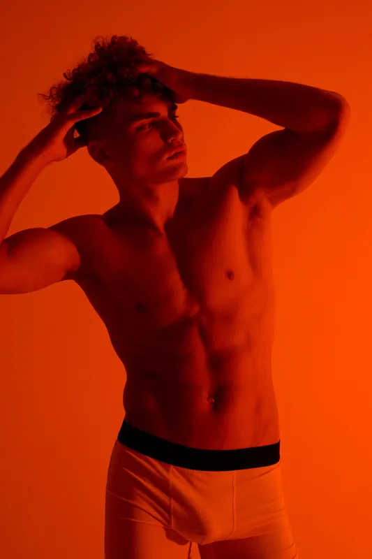 Young muscular man wearing light underwear posing with hands, showing defined abs and curly hair in orange studio lighting for fashion and fitness concept.
