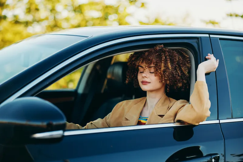 Woman driver in a vintage car wearing a tan blazer with curly hair, enjoying retro film color tones, relaxed smile, road trip mood, focused on road ahead
