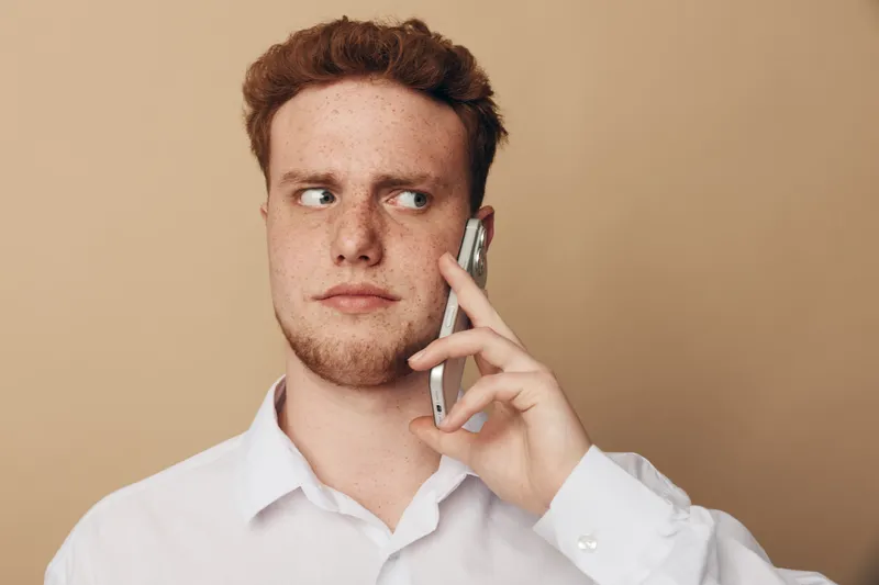 Young man with red hair and freckles wearing white shirt talks on smartphone looking sideways. Neutral beige background. Concept of communication, technology, and thoughtful expression.