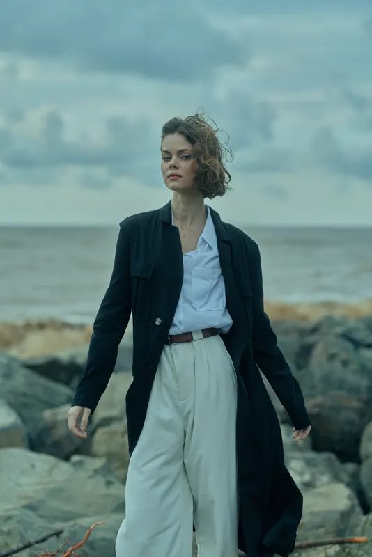 Woman walking along a rocky coastal line in a long dark coat and light trousers, wind in her hair, moody sea backdrop, outdoor fashion moment
