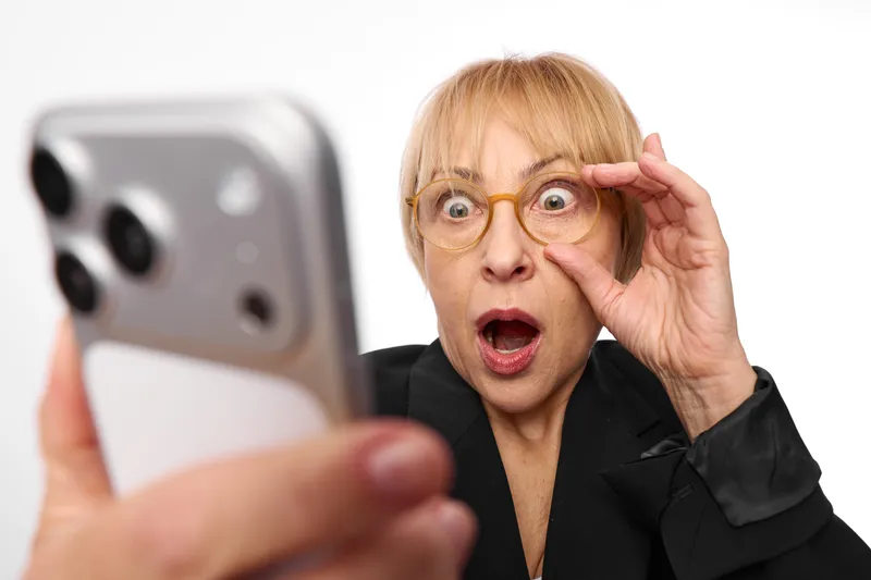 Senior woman, smartphone in hand, wears glasses, surprised expression, talking on video call, makes gesture near face, studio background, close up portrait