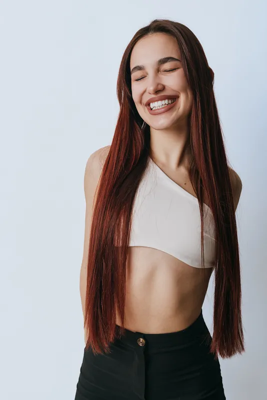 Young woman with long red hair smiling with closed eyes wearing white crop top and black pants standing against light background. Happy female portrait with natural beauty and casual style.