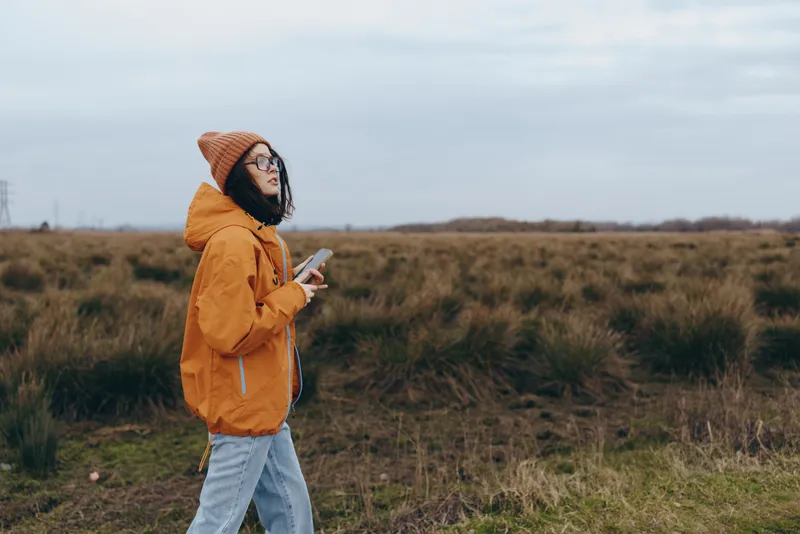 Young woman lifestyle outdoors, orange hoodie and backpack, walking in a grassy field while looking at smartphone, cloudy sky and wide nature landscape, casual travel mood, side view.