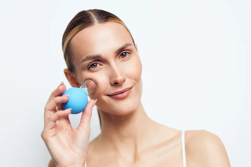 Young woman with light makeup holds a blue egg near her cheek, smiling softly in a bright studio with clean white backdrop, creating a fresh skincare moment