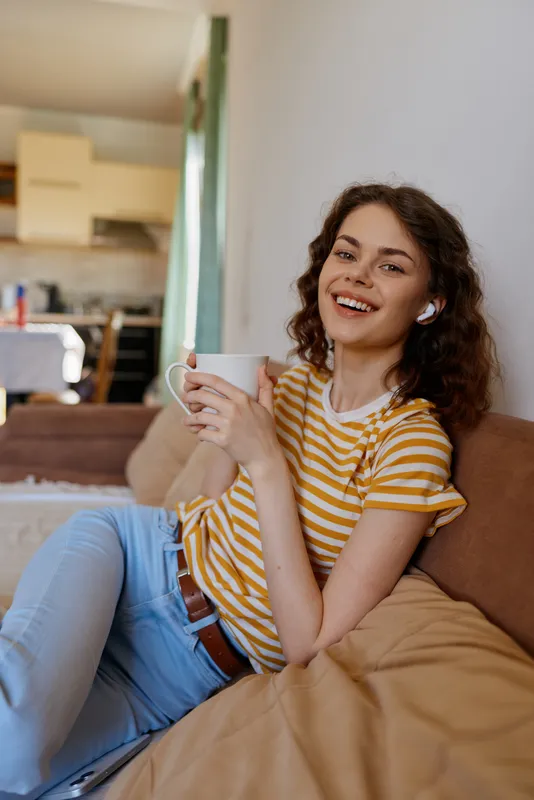 Smiling woman in a striped shirt sits on a sofa at home, holding a mug. Casual lifestyle moment with warm natural light and a cozy living room vibe, creating a welcoming mood.