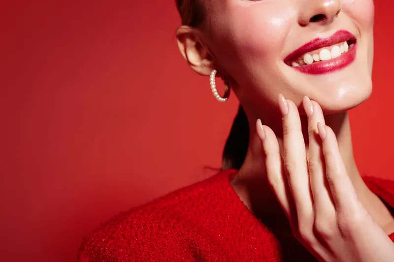 Close-up of smiling woman with red lipstick and pearl earring touching her face on red background. Concept of beauty, makeup, fashion, and happiness with vibrant studio lighting.