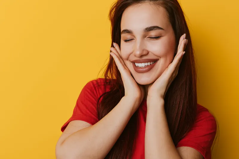 Woman smiling with eyes closed and hands on cheeks in red shirt against yellow background. Portrait of happy female with long hair enjoying moment in studio light.