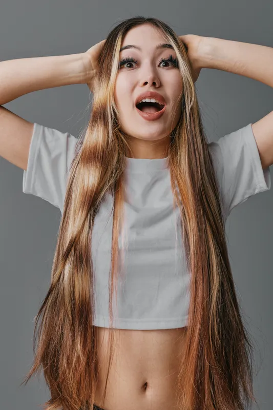 Young woman with long hair and braids, surprised expression, wearing white crop top, posing with hands on head against gray background indoors.