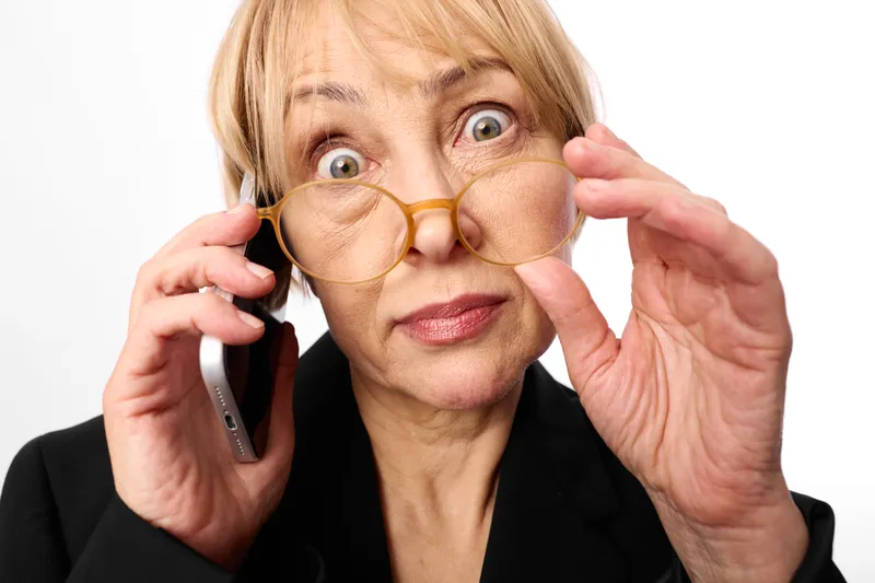 elderly woman glasses smartphone surprised expression, calling on mobile phone, closeup portrait, studio white background, thoughtful gesture, warm neutral colors, ready to help