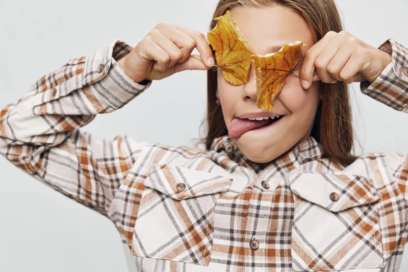 Child girl holding yellow autumn leaves in front of eyes playing funny face with tongue out casual checkered shirt on white background indoors.