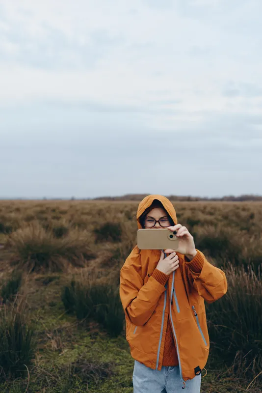 Woman outdoor lifestyle in grassy field, wearing orange jacket and hat, holding tablet for touch moment, looking at screen, cloudy sky, calm expression, nature colors, soft breeze
