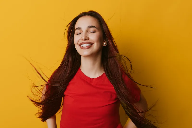 Young woman with long hair and closed eyes smiling happily against yellow background. Female in red shirt enjoying positive emotions with natural beauty and relaxed posture.