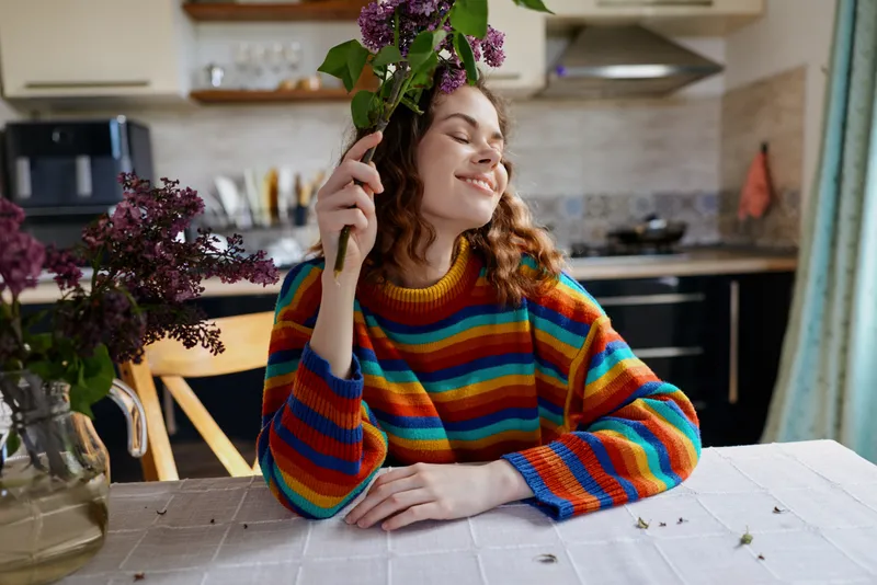 Smiling woman with colorful striped sweater sits at a kitchen table, holding purple flowers. Natural light creates a warm, cozy home mood and a relaxed lifestyle vibe.