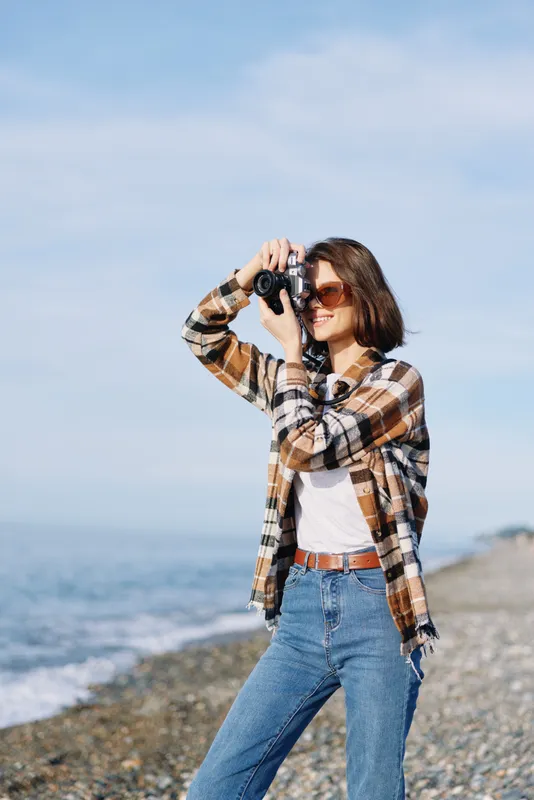 Photographer, woman, camera, beach, ocean, outdoor, photography, casual, plaid, sunglasses scene captures coastal mood with sunny weather and relaxed stance along a pebbled shoreline