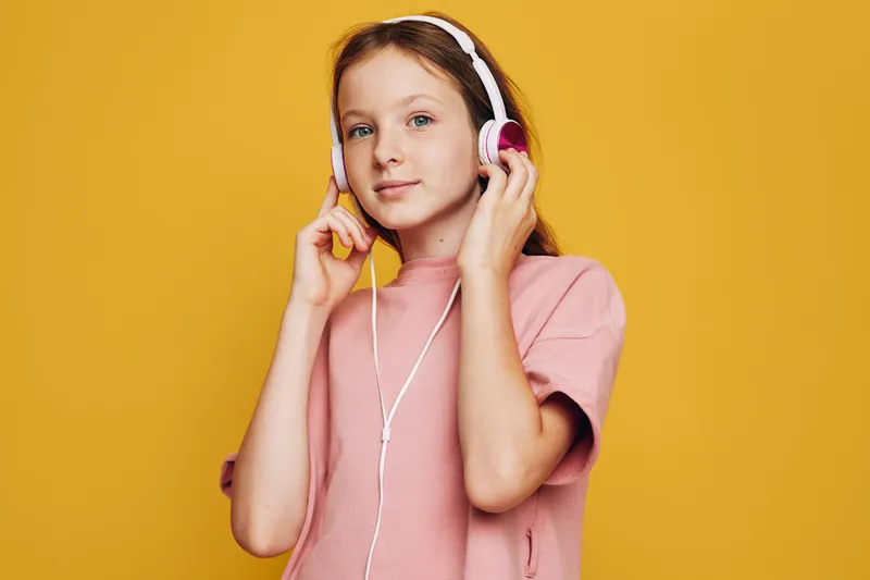 Young girl with red hair in pink t-shirt listens to music using white headphones on yellow background. Studio portrait with soft lighting and joyful mood.