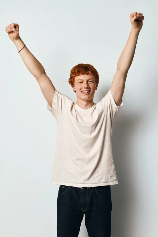 Young man with red hair and casual shirt raising arms in victory or celebration with smile on white background. Happiness, success and positive emotions in studio portrait.