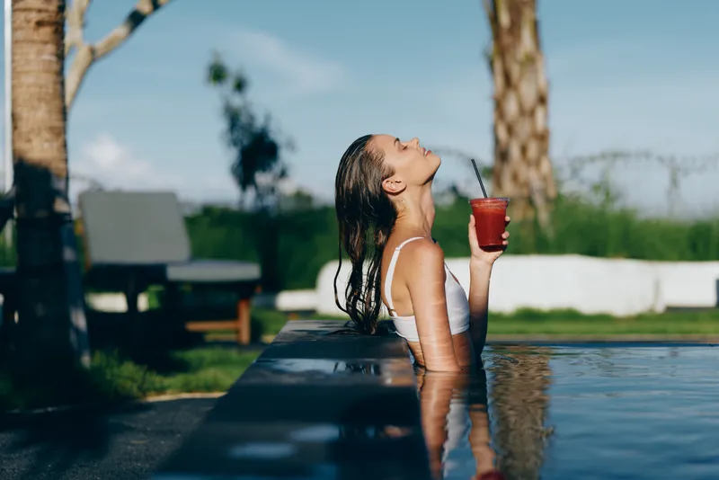 Young woman with braided hair relaxes in an outdoor pool holding a red drink with a straw while leaning back against the edge on a sunny day.