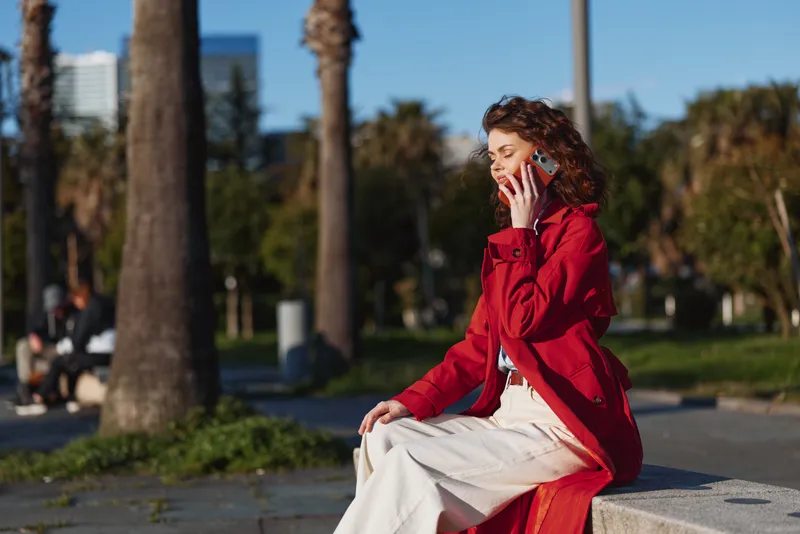 Young woman with curly hair wearing a red jacket talks on a smartphone while sitting on a bench in a sunny urban park with palm trees in the background.