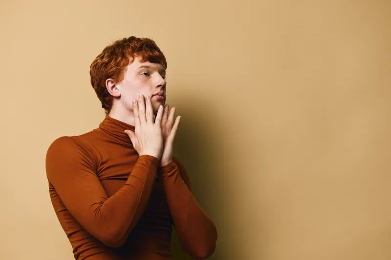 Young man with red hair wearing brown turtleneck sweater posing with hands near face on beige background. Portrait, thoughtful expression, fashion and style concept with copy space.