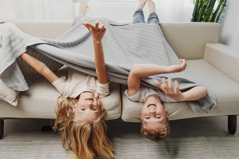 Two happy children lying upside down on beige couch covered with grey blanket, playing and smiling in cozy living room with casual home atmosphere.